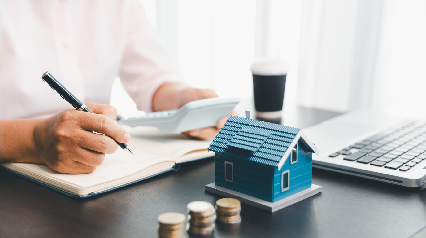 Person calculating mortgage costs with miniature house model coins notebook and laptop - home loan planning and financial budgeting workspace